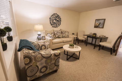 A cozy living room with a beige couch adorned with patterned cushions, a matching patterned armchair with a teal throw, a round coffee table with a small plant, a side table with a lamp and picture frame, a small dining table with two chairs, and a framed painting on the wall.