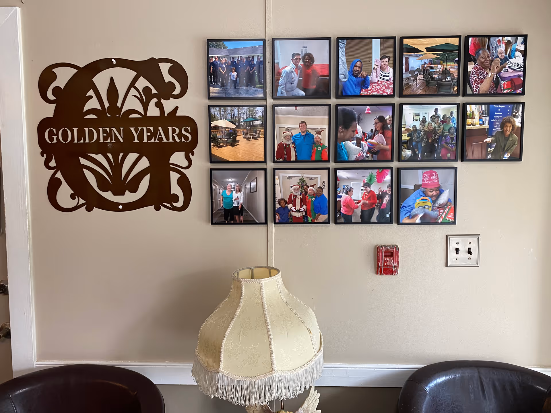 Interior wall of Golden Years Assisted Living Facility with a decorative metal sign reading 'GOLDEN YEARS' and a collage of framed photos showing residents and staff in various activities and social events. Below the photos is a vintage-style lamp with a fringed shade, flanked by two dark leather chairs. A fire alarm and electrical outlets are visible on the wall.