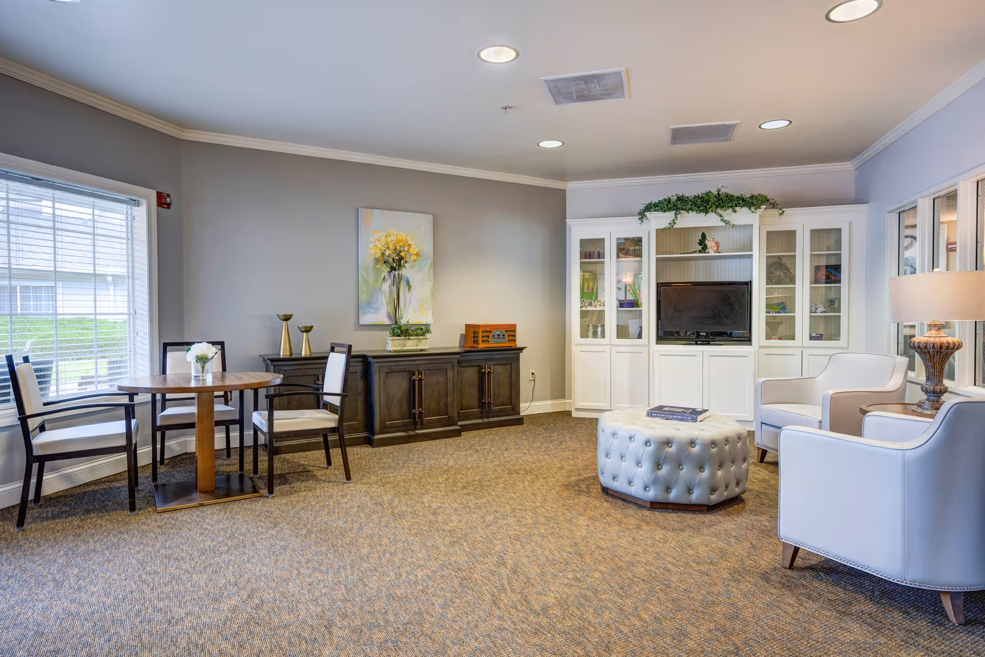A cozy living room area with a round wooden table and four chairs near a window with blinds. Against the wall is a dark wooden cabinet with decorative items and a painting of yellow flowers above it. On the right side, there is a white entertainment center with a TV and shelves holding various decorations. In front of the entertainment center is a tufted round ottoman, and two white armchairs with a lamp on a side table are positioned nearby. The room has beige carpet and neutral-colored walls.