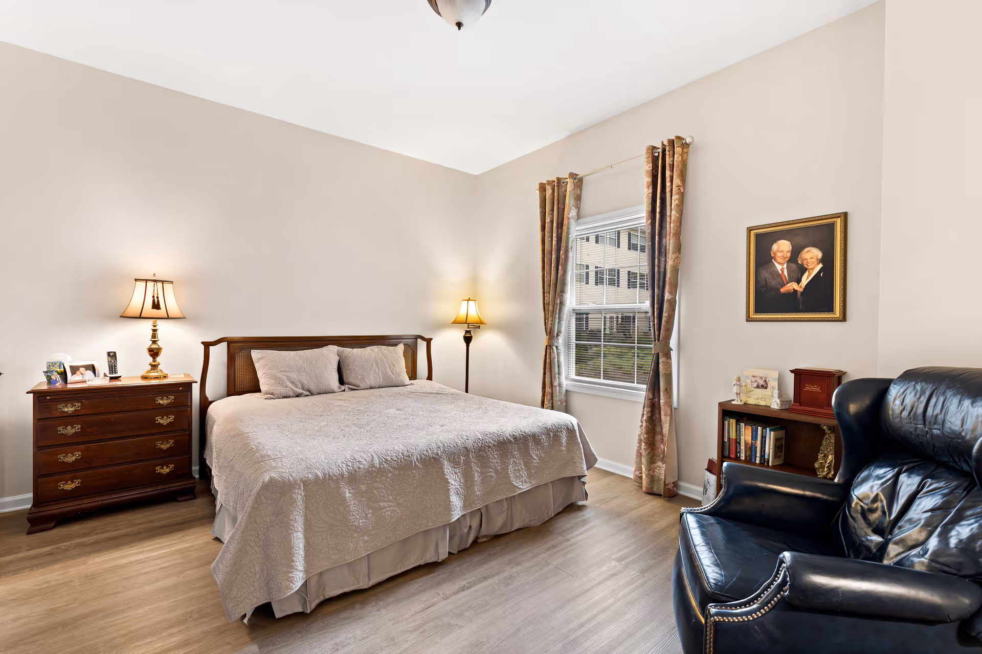A furnished bedroom with a bed, bedside tables and lamps, a window with curtains, an armchair and a framed portrait on the wall.