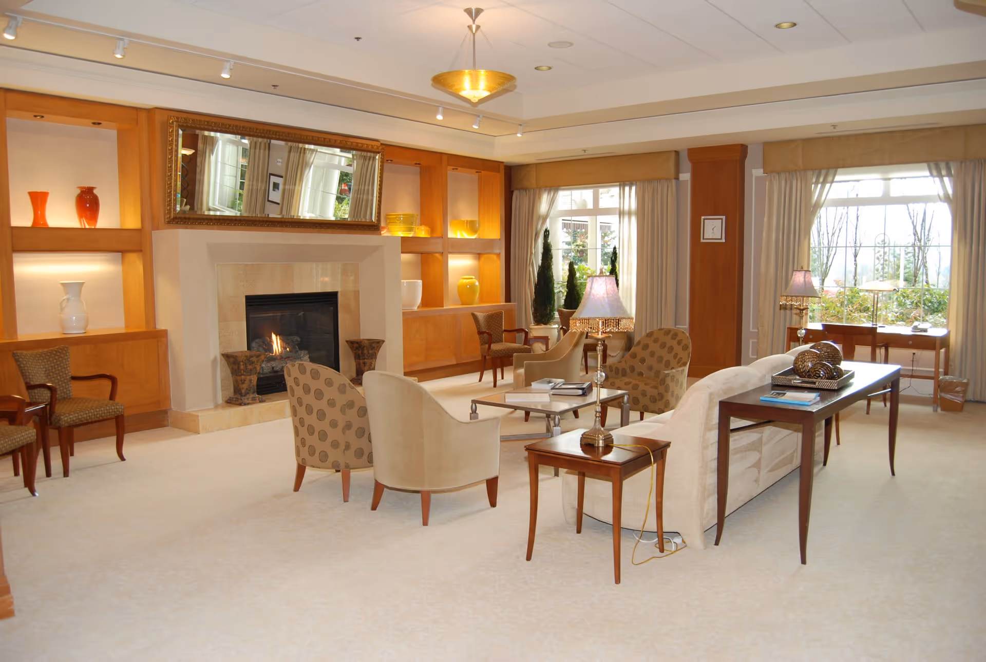 A spacious and well-lit living room with beige carpeting, a fireplace surrounded by a wooden built-in shelving unit displaying decorative vases, a large mirror above the fireplace, multiple armchairs and a sofa arranged around a coffee table, side tables with lamps, and large windows with curtains allowing natural light to fill the room.