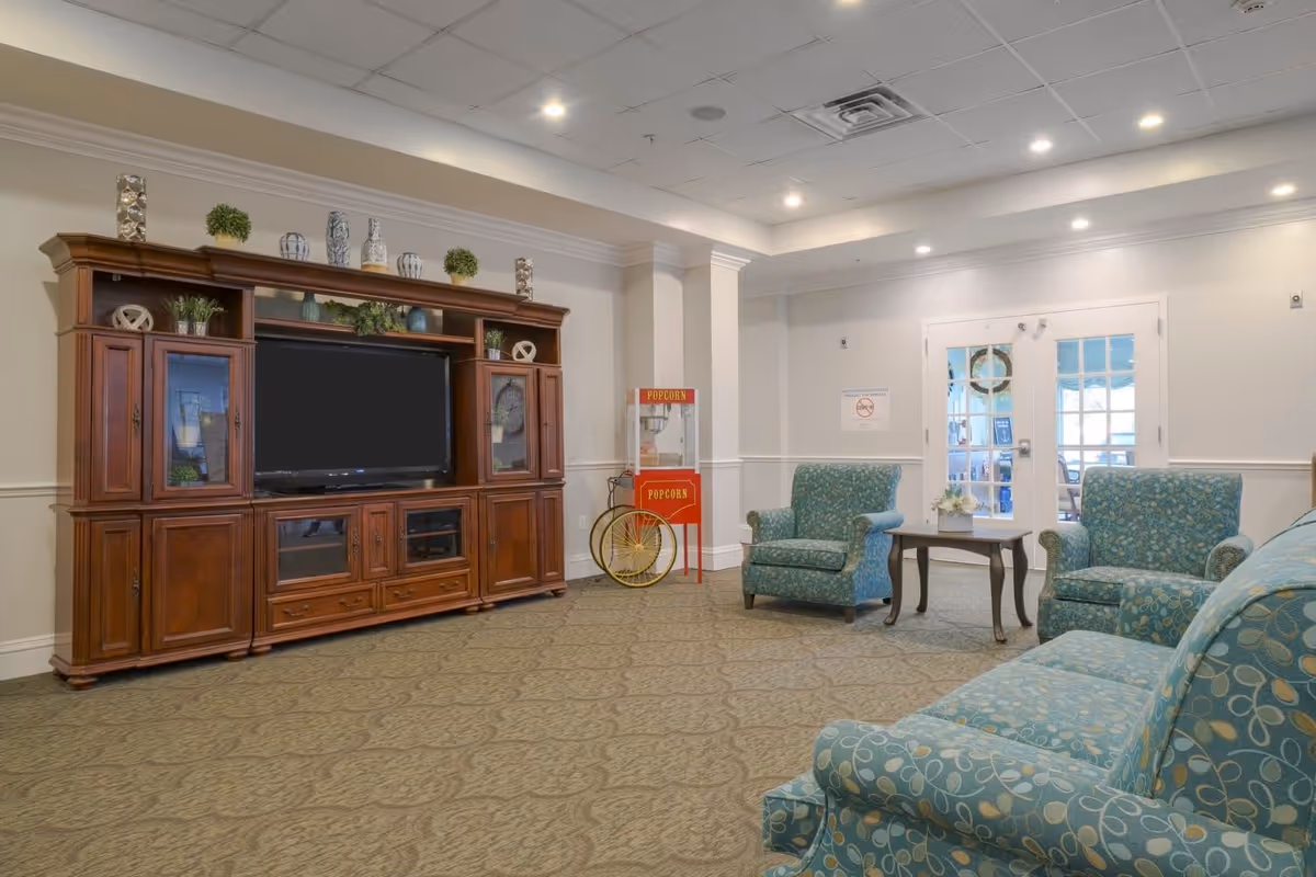 A senior living facility common area with a large wooden entertainment center holding a flat-screen TV, decorative plants, and vases. There are three upholstered armchairs and a sofa with blue patterned fabric arranged around a small wooden coffee table. A vintage-style popcorn machine is positioned near double glass doors, and the room has recessed lighting and a patterned carpet.