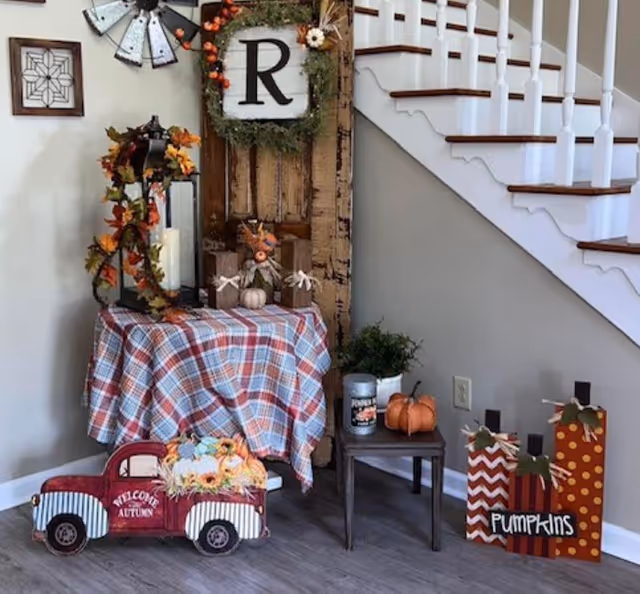 A cozy autumn-themed corner inside a home or facility with a small table covered in a plaid cloth, decorated with a lantern wrapped in fall leaves, small pumpkins, and rustic wooden decor. A wooden door with a wreath featuring the letter 'R' hangs on the wall behind the table. To the right, there is a small side table with a candle and a pumpkin, and three wooden pumpkin-shaped decorations with the word 'Pumpkins' written on one. A staircase with white railings and wooden steps is visible in the background.