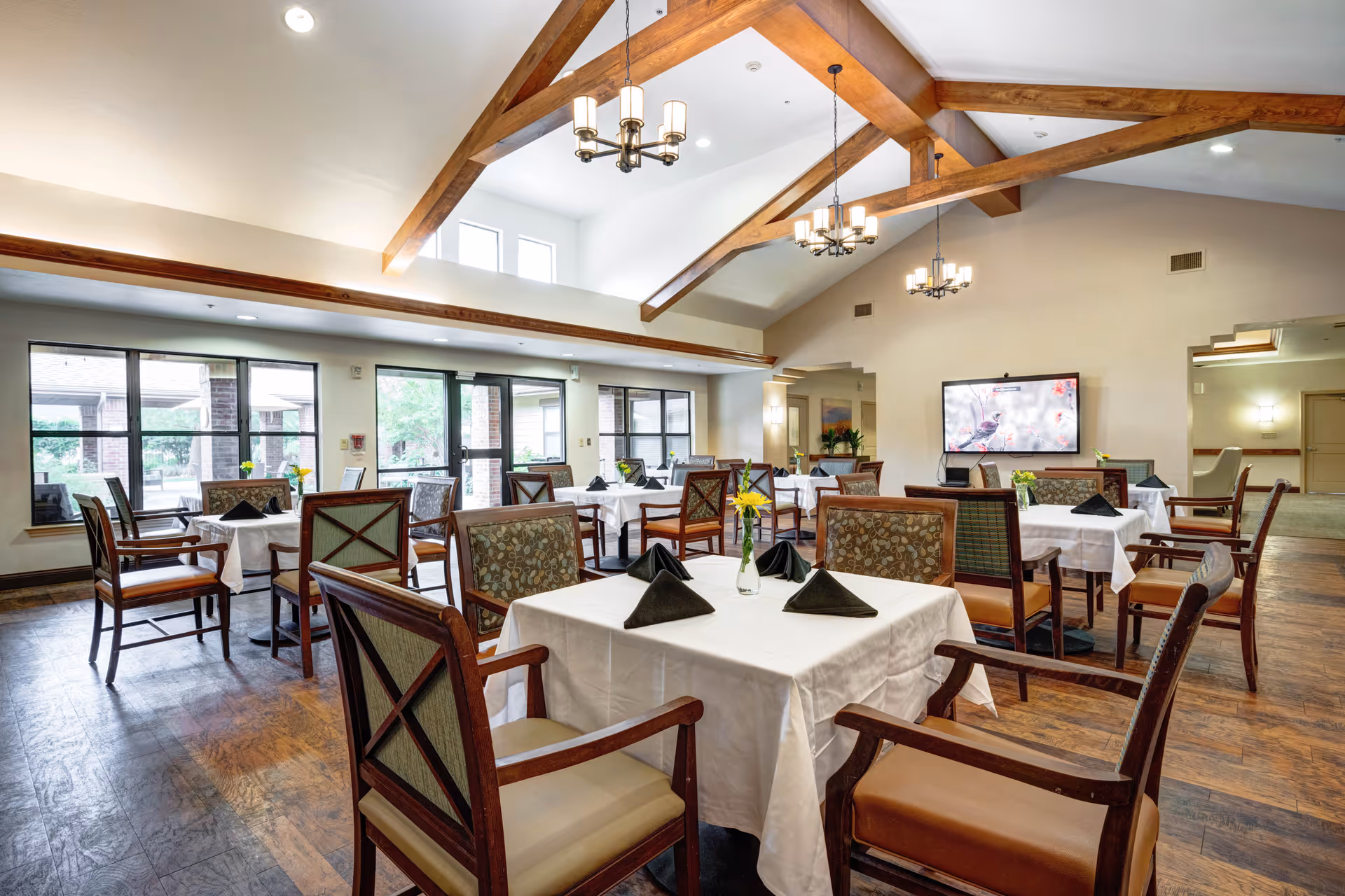 A spacious dining room with multiple tables covered in white tablecloths, each set with black folded napkins and small vases with yellow flowers. The room features wooden beams on the ceiling, chandeliers, large windows, and glass doors allowing natural light to enter. The floor is wooden, and there is a large flat-screen TV mounted on the far wall.