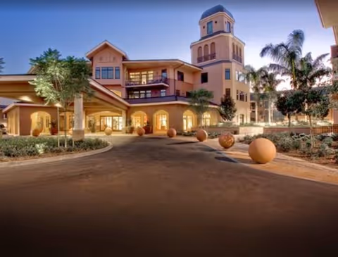 Exterior view of Walnut Village Orange County Retirement Community building at dusk, featuring a multi-story structure with a tower, arched windows, palm trees, and spherical decorative elements along the driveway.