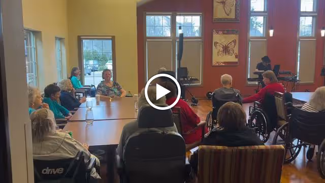 A group of elderly people seated around tables in a well-lit room with large windows and colorful walls, some in wheelchairs, engaged in an activity or meeting.
