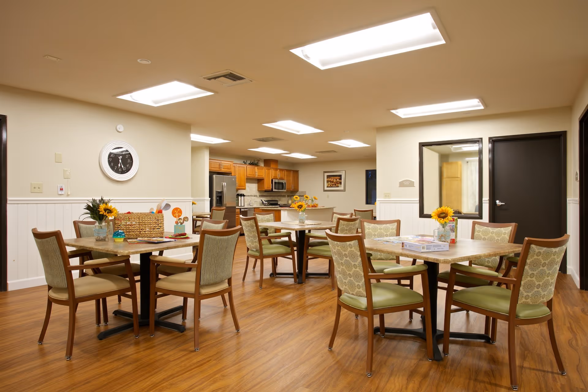 Bright communal dining room with several tables and chairs, sunflower centerpieces, and a kitchen area in the background.