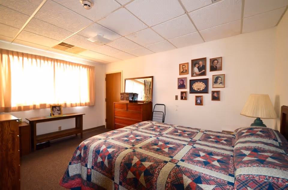 A modest furnished bedroom with a quilted bed, wooden dressers, a lamp, and a wall of framed photos beside a curtained window.