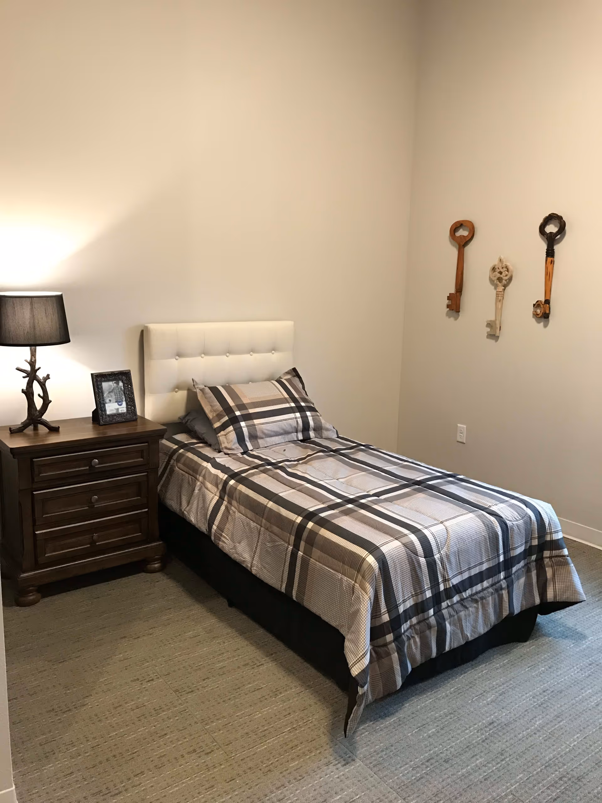 Small bedroom with a single bed dressed in plaid bedding, a wooden nightstand with a lamp and picture frame, and decorative oversized keys on the wall.