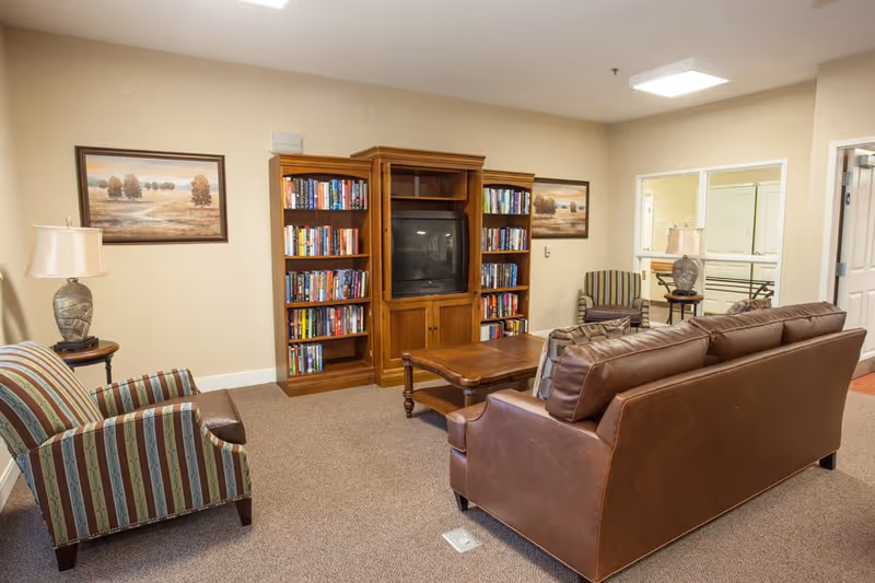 Cozy common living room with a leather sofa, striped armchairs, wooden bookcases filled with books and a TV, and a coffee table.