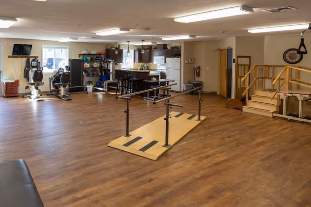 A spacious room with wooden flooring featuring parallel bars for physical therapy in the center. To the left, there are exercise machines and shelves with various fitness equipment. In the background, there is a kitchenette area with a refrigerator, cabinets, and a small dining table with chairs. On the right side, there is a set of wooden stairs with handrails and a clock on the wall.