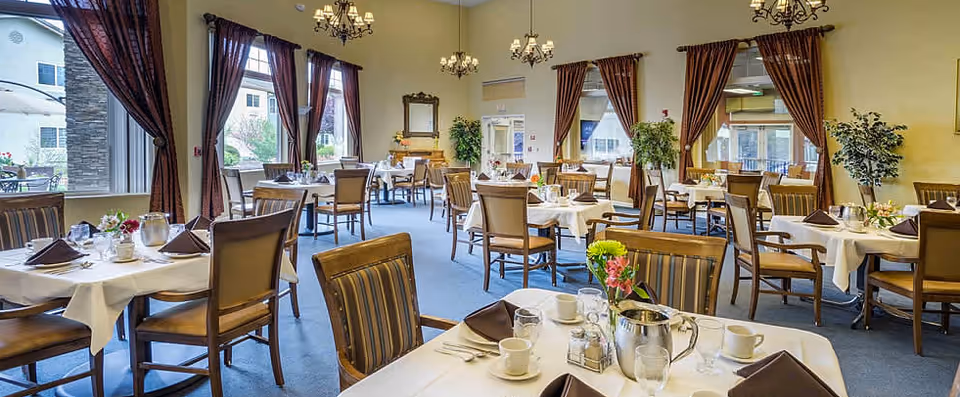 A spacious dining room with multiple tables covered in white tablecloths, each set with cups, glasses, napkins, and small flower arrangements. The room features large windows with brown curtains, chandeliers hanging from the ceiling, and several plants placed around the room. The atmosphere is bright and inviting.