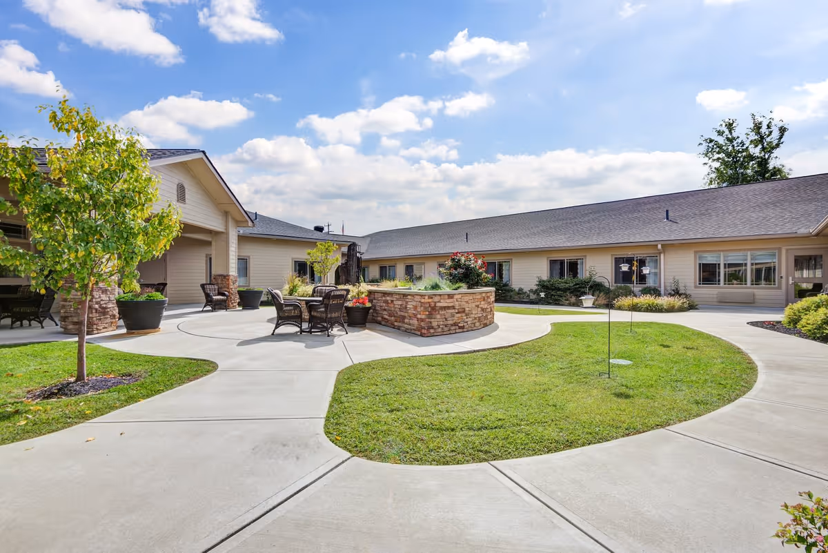 Outdoor courtyard area of Juniper Glen Memory Care Assisted Living with a circular concrete walkway surrounding a grassy area with a raised brick planter in the center. Several chairs and tables are placed on the walkway near the building. The building is single-story with beige siding and multiple windows. The sky is partly cloudy with blue patches.