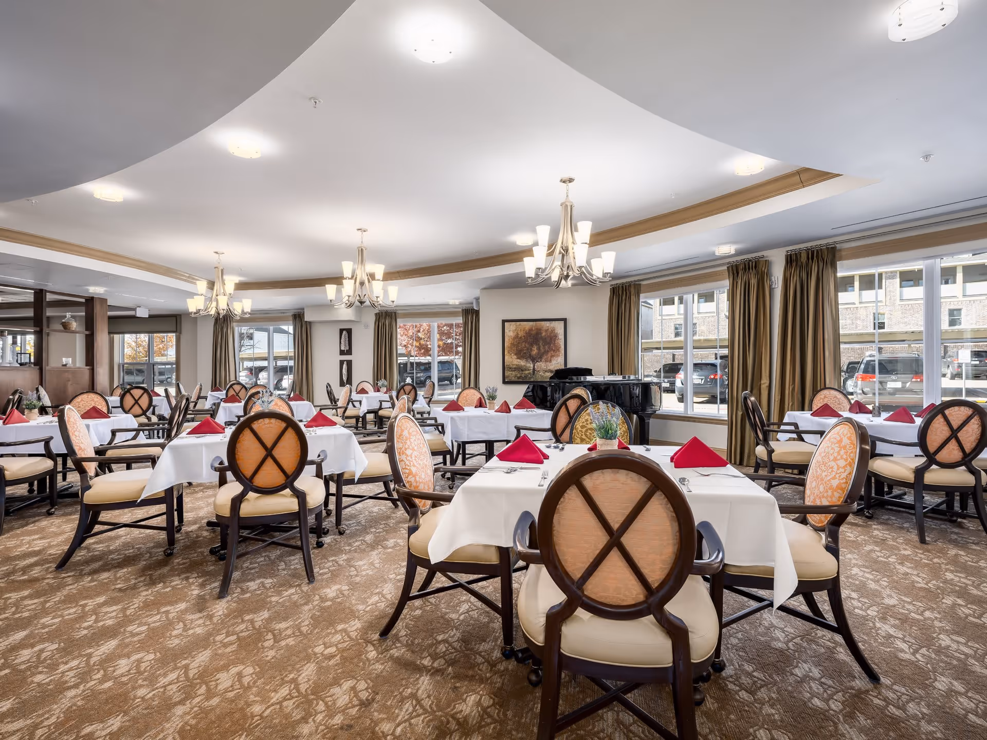 A spacious dining room with multiple tables covered in white tablecloths, each set with red folded napkins and silverware. The room features elegant chairs with cushioned seats and backs, large windows with beige curtains allowing natural light, chandeliers hanging from the ceiling, and a grand piano in the background.
