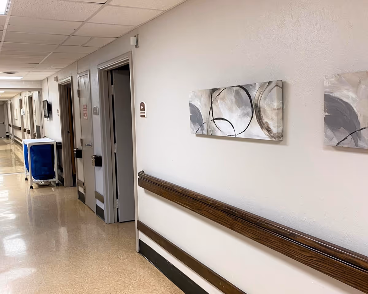 A clean, well-lit hallway in a care facility with beige walls and floor, wooden handrails, several closed doors, and abstract artwork on the wall. A blue cart is visible further down the corridor.
