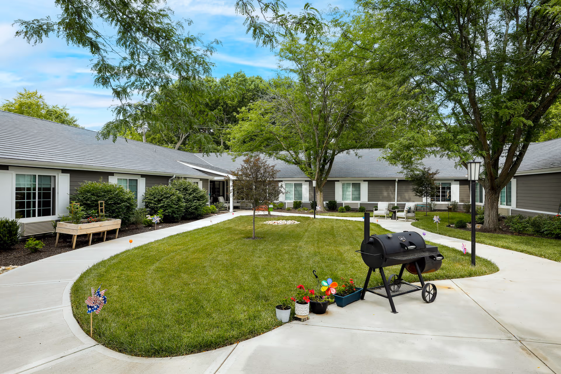 A courtyard area at Homestead Estates Assisted Living of Leawood featuring a well-maintained grassy lawn surrounded by a paved walkway. There are several trees providing shade, a black outdoor grill, potted plants, and some outdoor seating near the building. The building has gray siding with white trim and multiple windows.