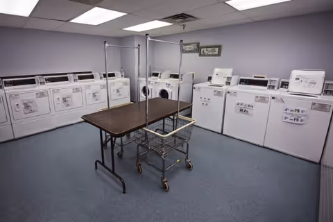 Laundry room with multiple white washing machines and dryers lined up against the walls. In the center, there is a folding table and a laundry cart with a metal frame and wheels. The walls are painted light purple and the floor is covered with blue linoleum tiles. Two framed pictures hang on the wall above the machines.