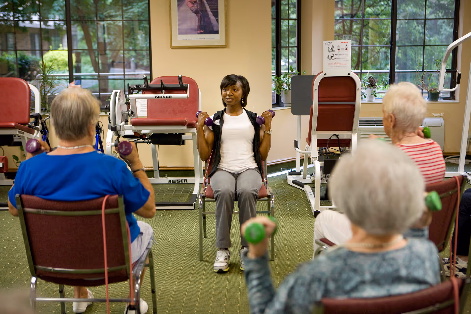 A fitness instructor leads a seated exercise class for seniors in a room with large windows and exercise equipment. The seniors are sitting in chairs and holding small dumbbells while following the instructor's movements.