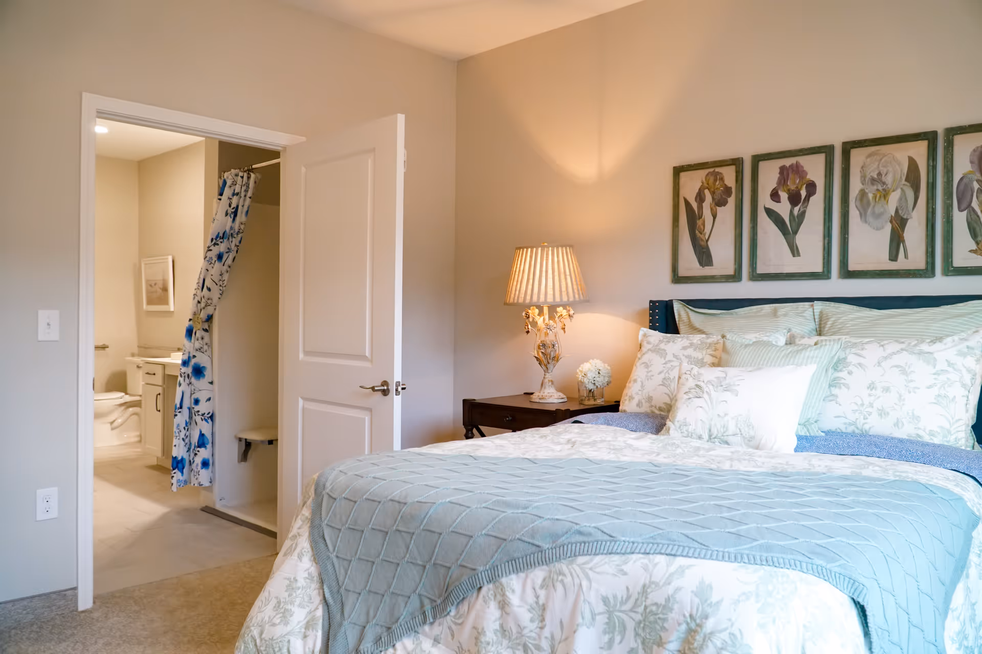 A cozy bedroom with a neatly made bed featuring floral and striped pillows and a light blue quilt. A bedside table with a decorative lamp and a small flower arrangement is next to the bed. Above the bed are five framed botanical prints. An open door reveals an adjacent bathroom with a shower curtain decorated with blue flowers.
