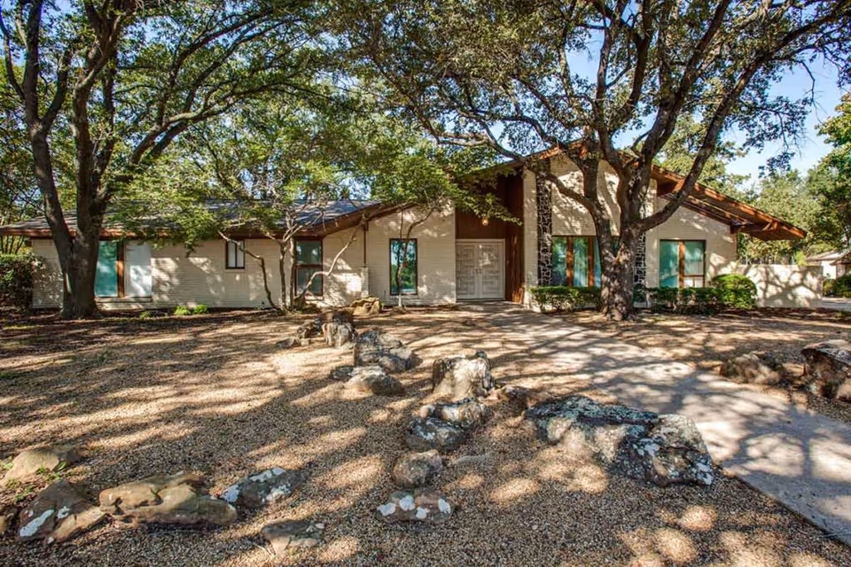 Exterior view of a single-story building with a sloped roof, surrounded by large trees casting shadows on the ground covered with gravel and rocks. The building has multiple windows and a central entrance door.