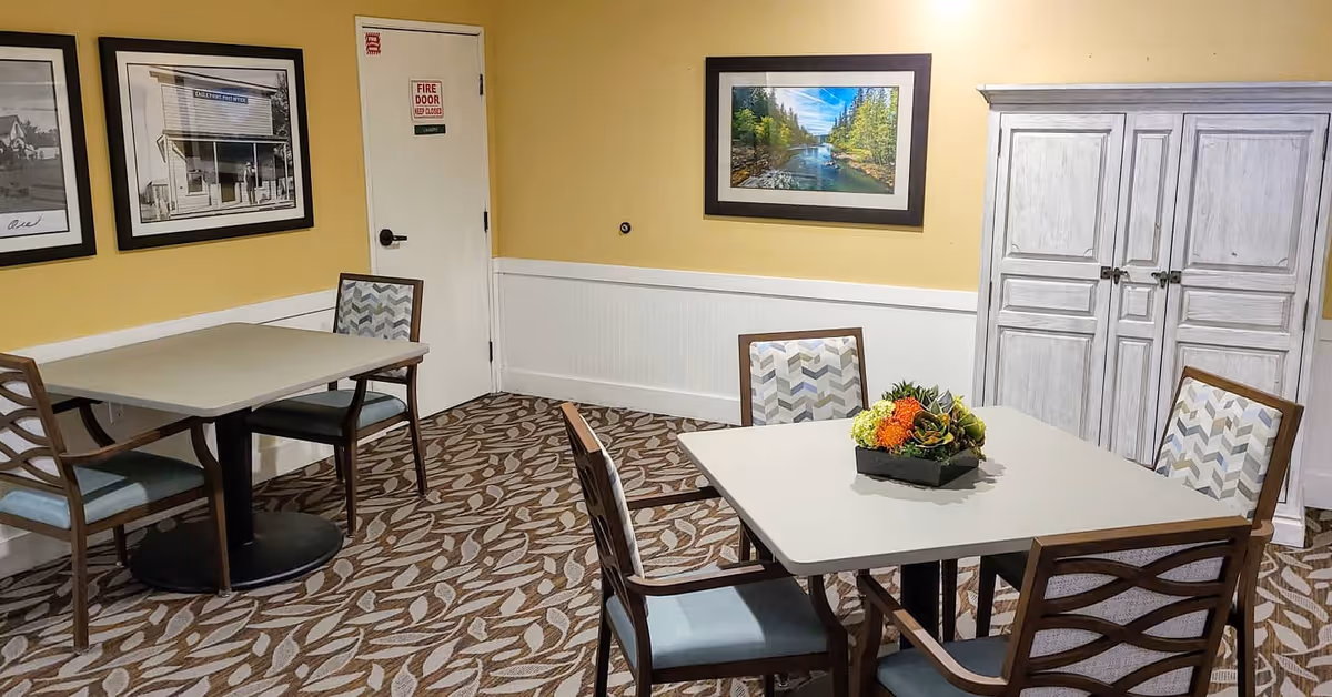 Small dining room with two square tables and chairs, patterned carpet, framed artwork on yellow walls, and a white storage cabinet.