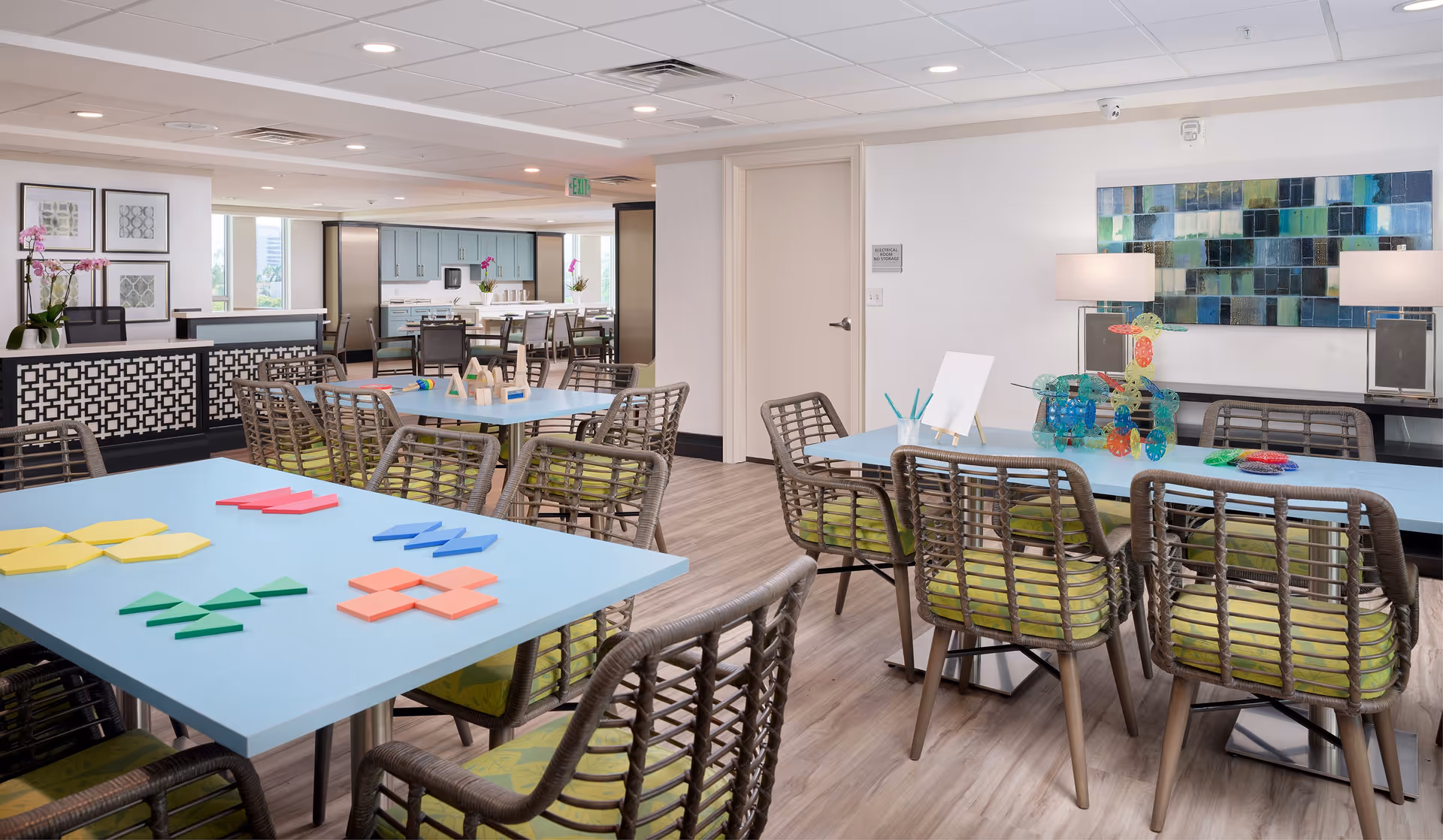 A bright and spacious activity room in a senior living facility with multiple tables and chairs. The tables have colorful geometric shapes and building toys on them. The room features light blue tabletops, wicker chairs with green cushions, wooden flooring, and modern decor including framed artwork and a large abstract painting on the wall. In the background, there is a kitchenette area with cabinets and a refrigerator.