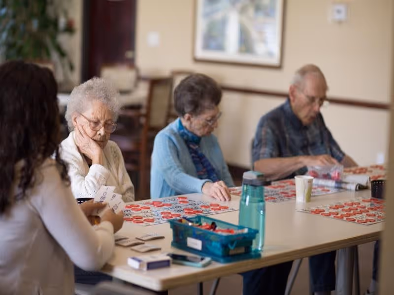 Three elderly individuals sitting at a table playing bingo with a younger person assisting them in a well-lit room with framed artwork on the wall.