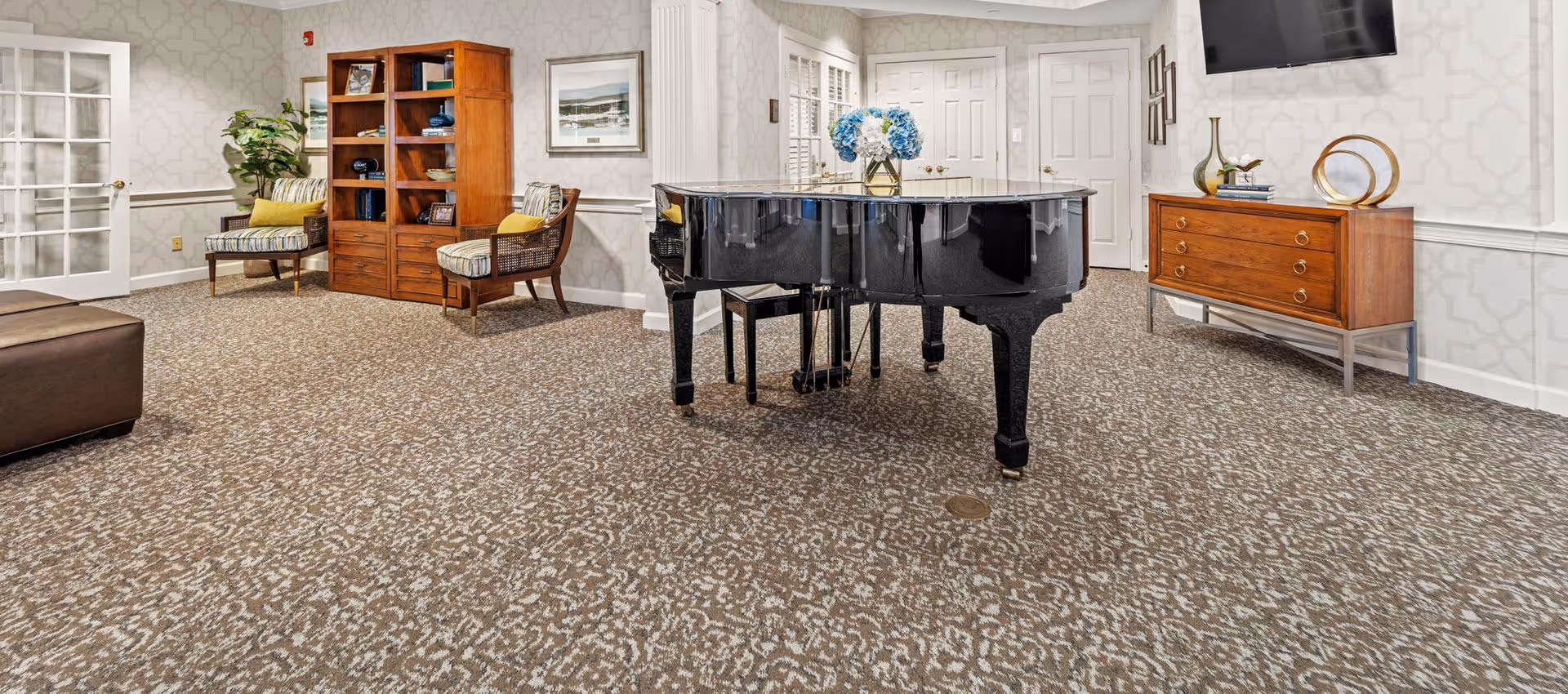 Carpeted communal lounge with a black grand piano, seating, wooden bookcase and dresser beneath a wall-mounted TV.