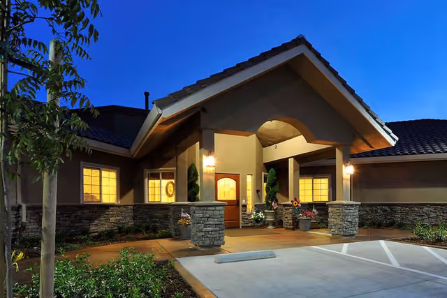 Exterior view of a senior living facility entrance at dusk with warm lights glowing from inside. The building has a covered entryway supported by stone pillars, a wooden door, and windows with yellow light. There are plants and flowers near the entrance and a parking area in front.