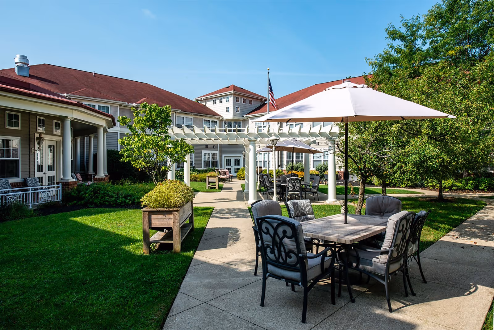Outdoor patio area at SouthWest Commons by New Perspective featuring tables with umbrellas and cushioned chairs on a paved walkway, surrounded by green grass, trees, and a building with red roofs in the background under a clear blue sky.