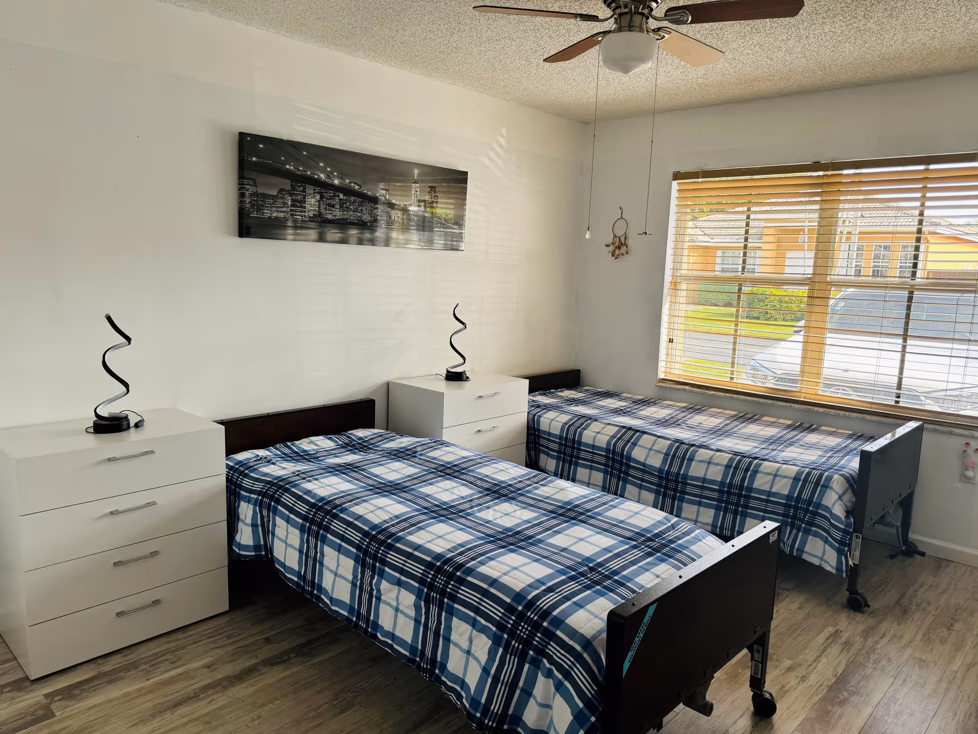 A bedroom with two single beds covered in blue and white plaid bedding. Each bed has a white nightstand beside it with a modern black spiral lamp on top. A large window with wooden blinds lets in natural light, and a ceiling fan with wooden blades is mounted on the ceiling. A black and white cityscape picture hangs on the wall above the beds.