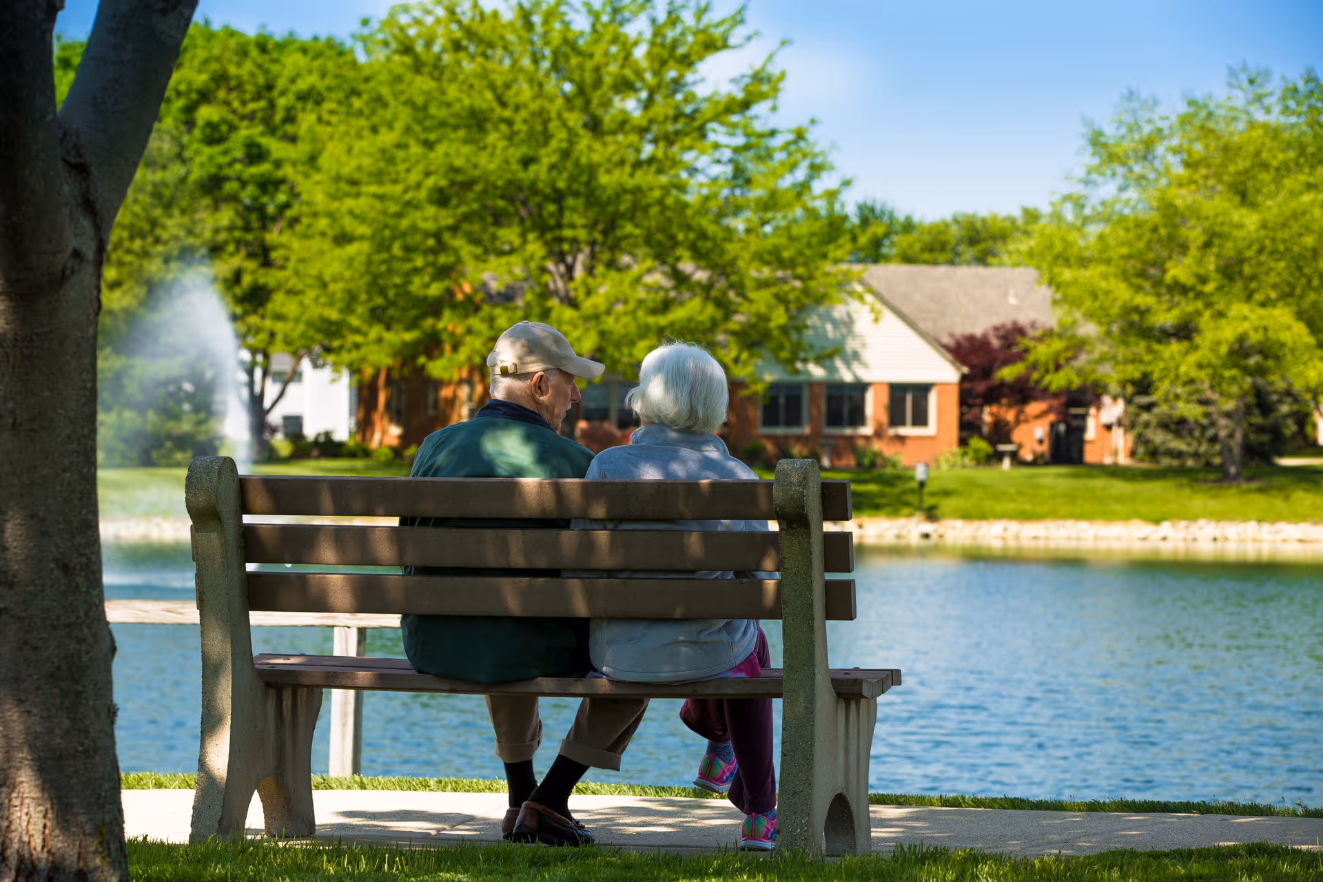 An elderly couple sitting on a wooden bench by a lake with a fountain, surrounded by green trees and residential buildings in the background on a sunny day.