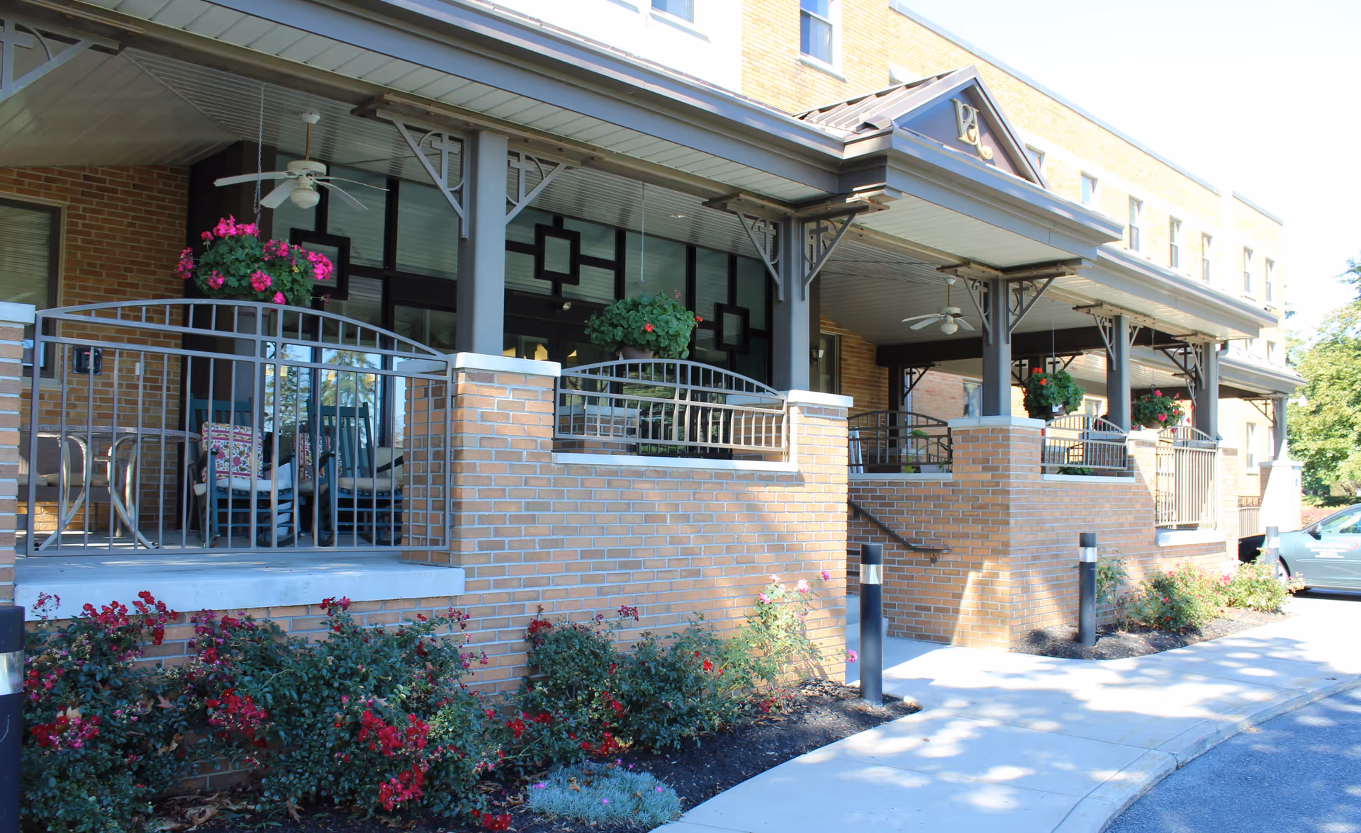 Covered brick front porch entrance with hanging flower baskets, metal railings, and seating at a retirement community building.