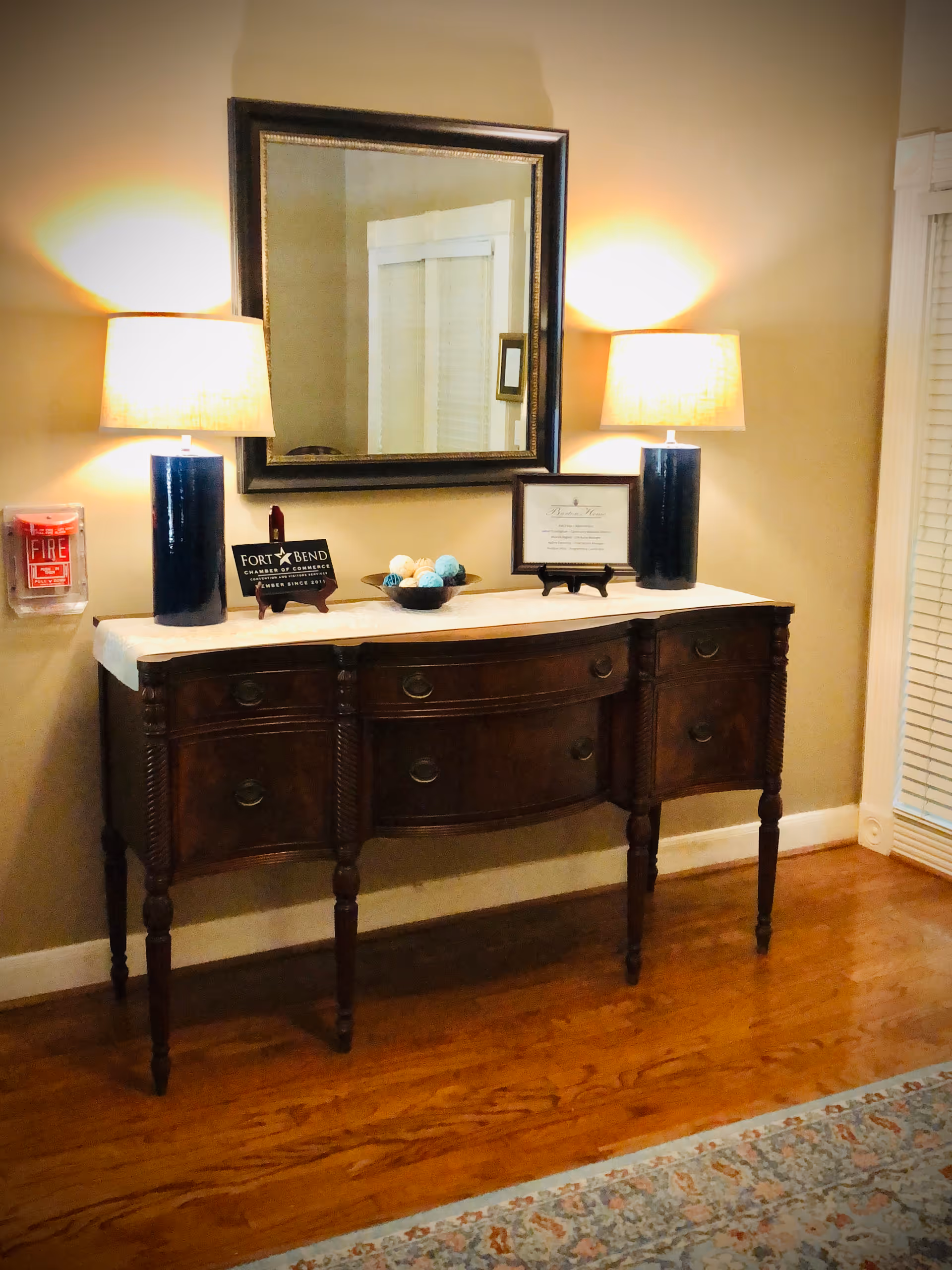 Wooden console table beneath a mirror topped with two lamps, a decorative bowl and plaques in an interior hallway.