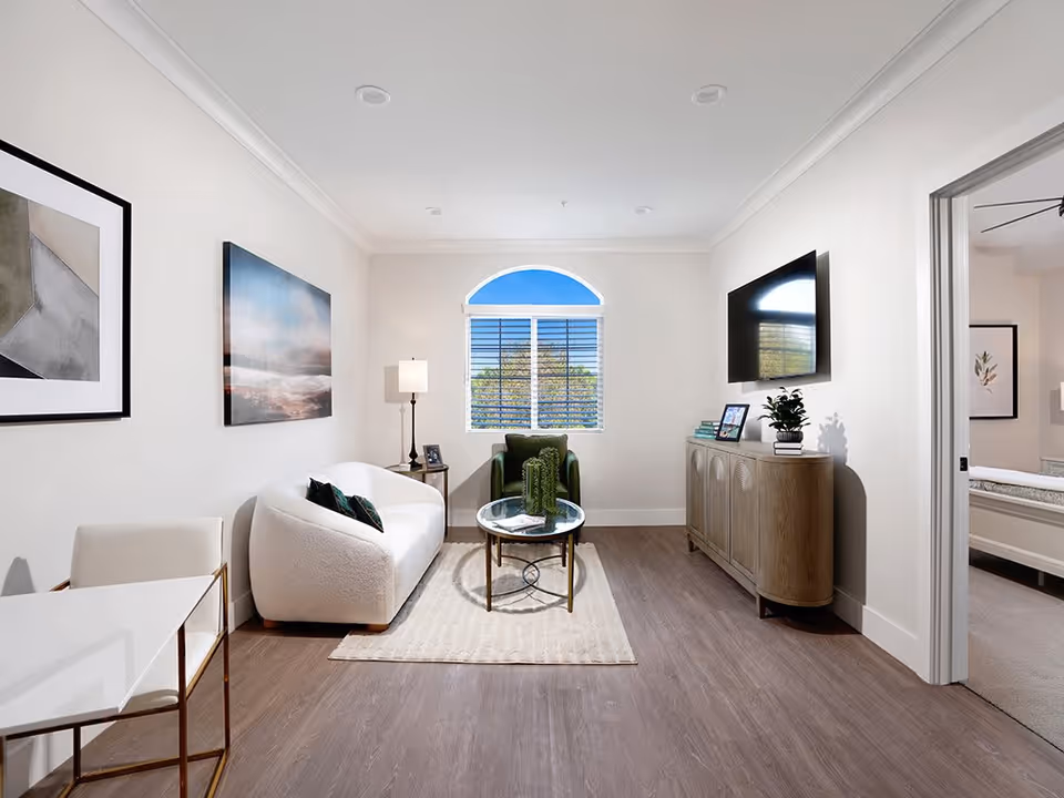 A bright and modern living room with a white curved sofa, a green armchair, a round glass coffee table on a beige rug, a wooden sideboard with a flat-screen TV mounted above it, and a window with white blinds and an arched top letting in natural light. There is a floor lamp next to the sofa and framed artwork on the walls. An open doorway leads to a bedroom with a bed and framed artwork visible.