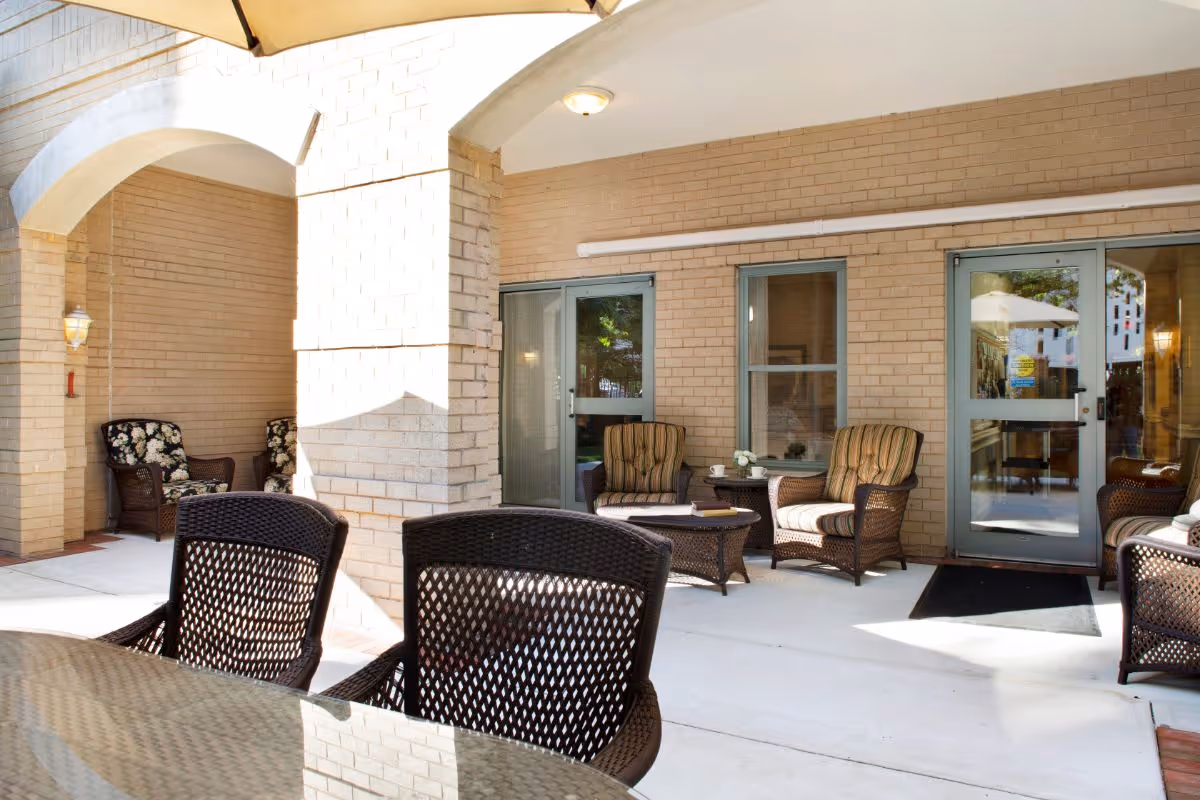 Covered outdoor patio with wicker chairs and tables outside a brick building entrance.