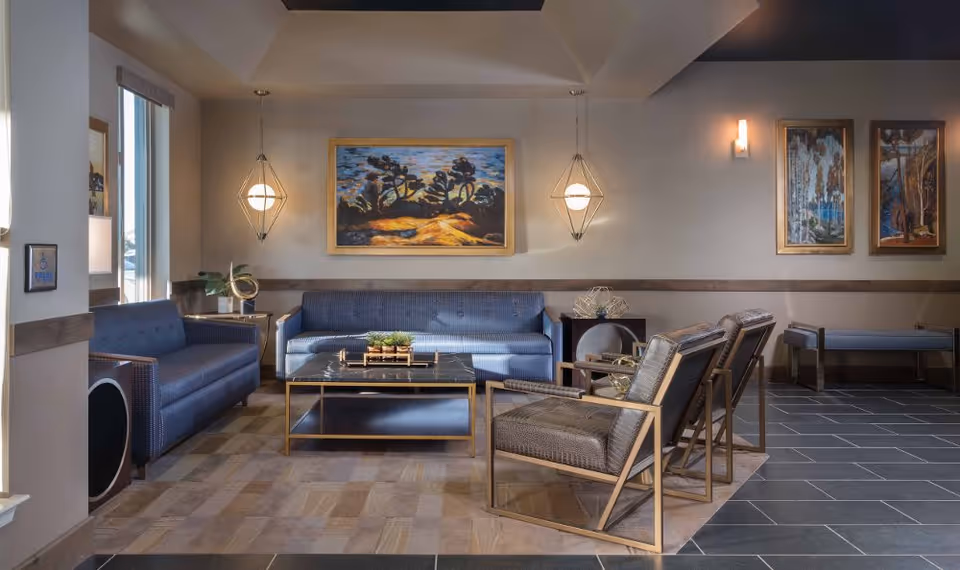 A modern and comfortable living room area with two blue sofas, two armchairs with gold frames, a black marble coffee table, and decorative wall art. The room is well-lit with hanging pendant lights and wall sconces, featuring a mix of tiled and carpeted flooring.