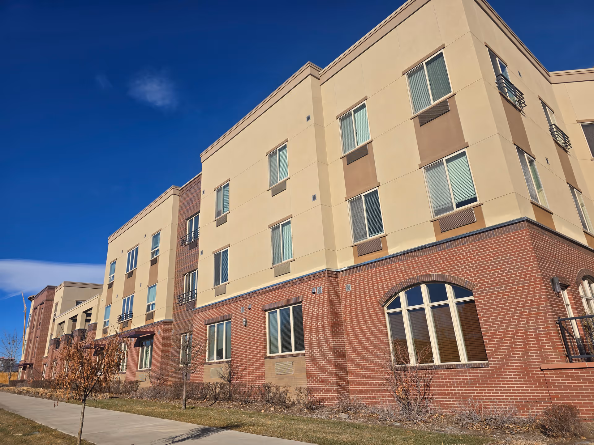 Facade of a three-story brick and stucco senior living building with many windows and a sidewalk in front under a clear blue sky.