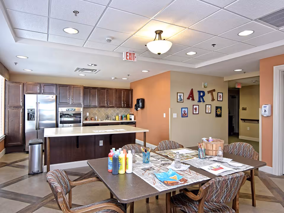A bright and spacious art activity room in a senior living facility featuring a large table with chairs around it. The table is set up with acrylic paint bottles, paintbrushes, water bottles, and art projects on newspaper. In the background, there is a kitchen area with dark wooden cabinets, a stainless steel refrigerator, and an oven. The wall has decorative letters spelling 'ART' along with framed artwork. The ceiling has recessed lighting and a hanging light fixture, and there is an exit sign visible.