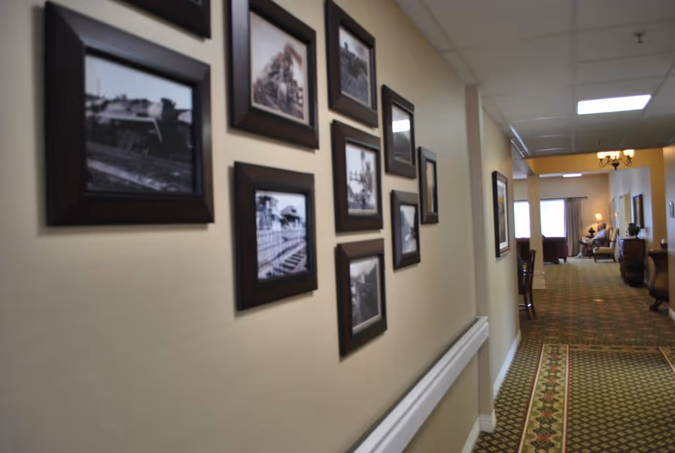 A hallway in a senior living facility with framed black and white photographs on the left wall. The hallway has patterned carpet and leads to a sitting area with chairs and a lamp near windows at the far end.