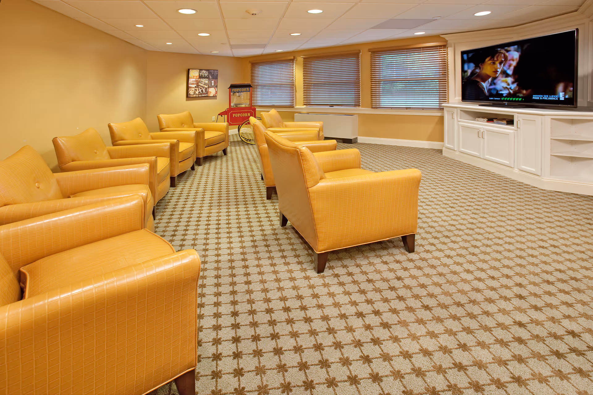 A cozy lounge area with multiple mustard-yellow armchairs arranged in two rows facing a large flat-screen TV mounted on a white built-in cabinet. The room has beige walls, patterned carpet flooring, three windows with blinds, and a vintage-style popcorn machine in the corner.