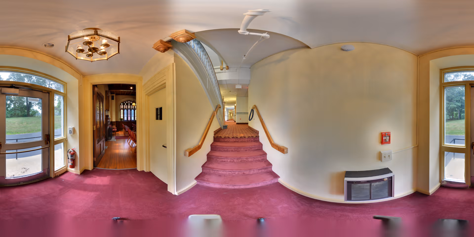Interior view of a senior living facility hallway with maroon carpet and cream-colored walls. There is a set of stairs with wooden handrails leading up to a corridor. To the left, there is a doorway leading into a room with wooden floors and chairs, and large windows letting in natural light. The hallway has large windows on both sides, allowing views of green outdoor areas. A fire extinguisher and a fire alarm are mounted on the walls.