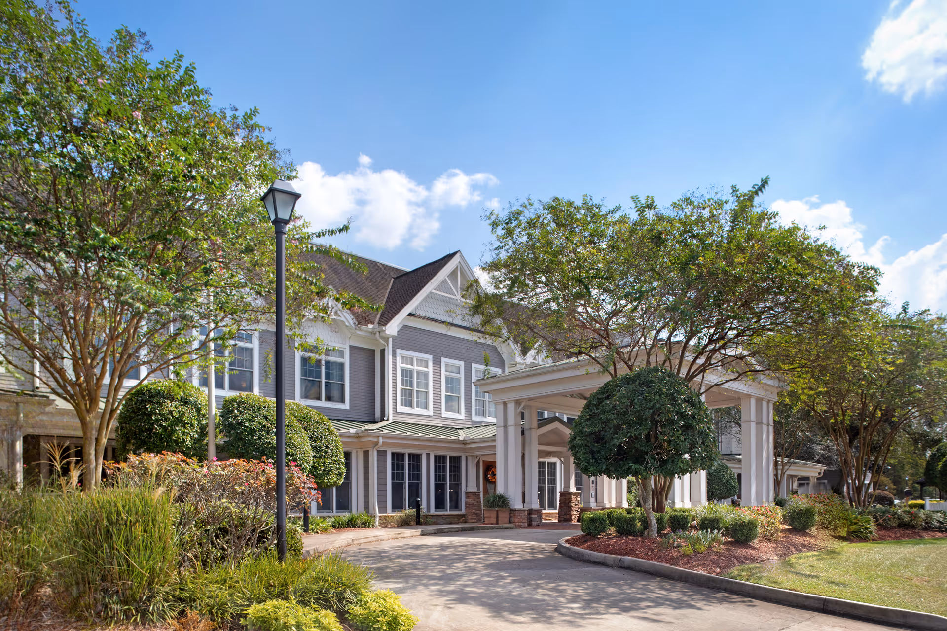 Front entrance of a two-story senior living building with a covered porte-cochere, landscaped driveway and trees.