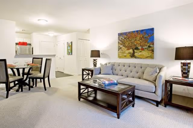 Bright furnished living room with a tufted sofa, coffee table, side tables, lamps, and a dining area with chairs visible near the kitchen.