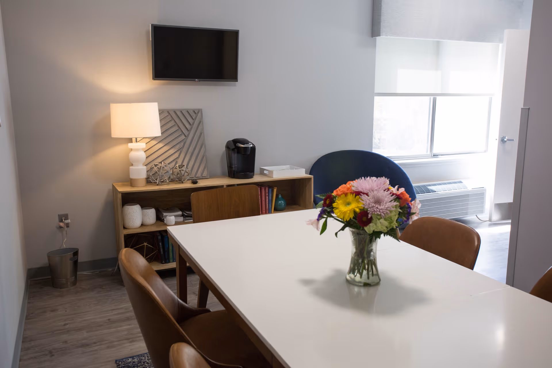 A small dining or meeting area with a white table surrounded by brown chairs. A vase with colorful flowers is placed on the table. In the background, there is a wooden cabinet with decorative items, a lamp, and a coffee maker. A flat-screen TV is mounted on the wall above the cabinet. A window with a roller shade lets in natural light, and an air conditioning unit is visible below the window.