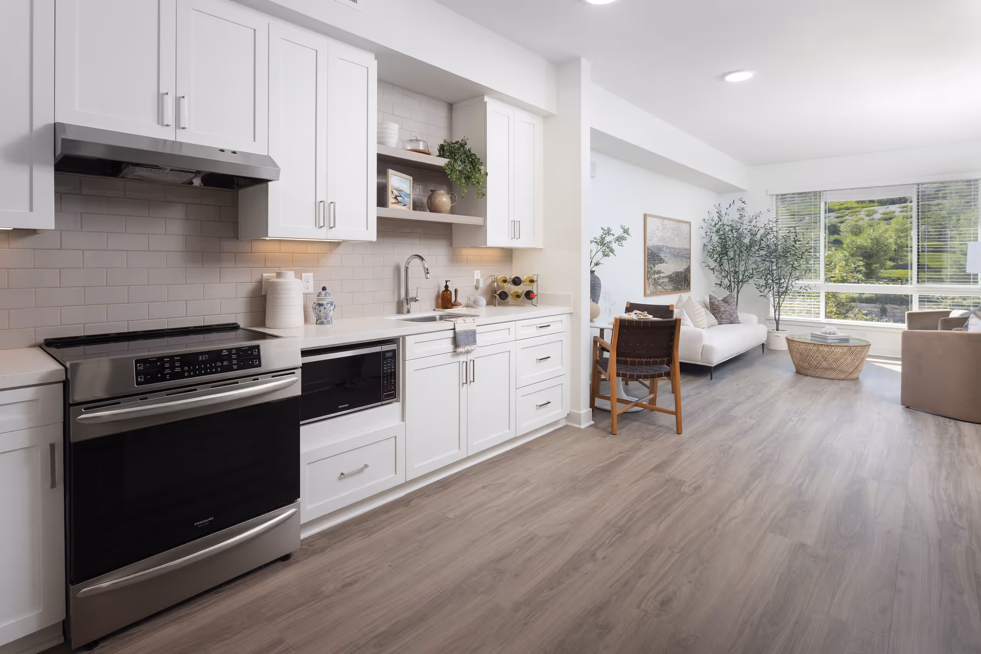 A modern open-concept kitchen and living area with white cabinetry, stainless steel appliances including an oven and microwave, a sink with a faucet, and decorative items on shelves. The living area has a white sofa, a wooden chair, a round wicker coffee table, large windows with blinds, and green plants, allowing natural light to fill the space.