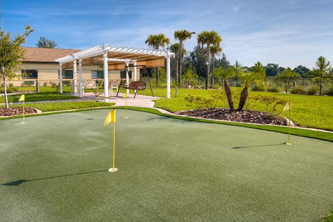 Outdoor putting green with several yellow flags in a landscaped area featuring green grass, palm trees, and a pergola with seating in the background under a clear blue sky.