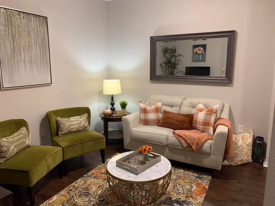 A cozy living room with a beige loveseat adorned with plaid and solid pillows and a rust-colored throw blanket. Two green upholstered chairs with patterned pillows are positioned to the left. A round coffee table with a marble top and gold metal base holds a small tray with a flower vase. A side table with a lamp, small plant, and decorative pumpkin sits between the loveseat and chairs. A large rectangular mirror hangs on the wall above the loveseat, reflecting a plant and floral artwork. The room features a patterned area rug and hardwood flooring.