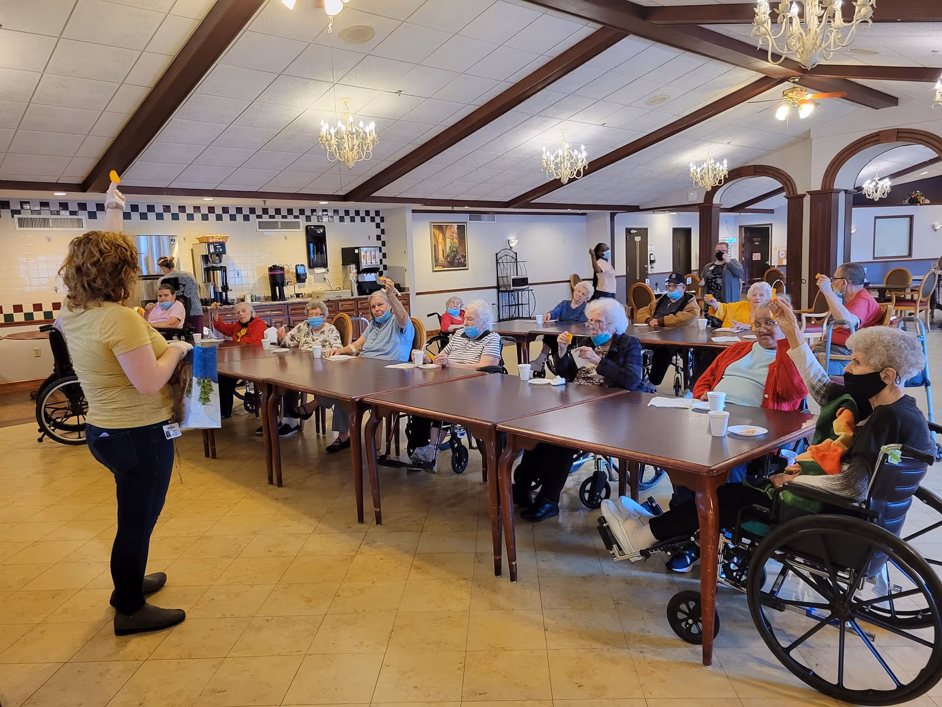 A group of elderly people sitting around tables in a dining or activity room, some in wheelchairs, raising their hands while a woman stands in front of them holding a bag. The room has chandeliers, a tiled wall with coffee machines, and a high ceiling with wooden beams.
