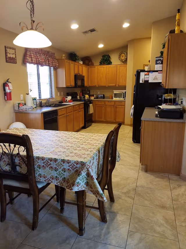 A kitchen area with wooden cabinets, a black refrigerator, a stove, a microwave, and a toaster oven. There is a dining table covered with a patterned tablecloth and surrounded by wooden chairs. The room is well-lit with ceiling lights and a hanging lamp above the table. A window with a red and white checkered valance is visible above the sink. Various kitchen items and decorations are placed on the countertops and cabinets.